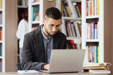 Man with eyeglasses wearing shirt and jacket working in library, browsing internet on laptop, reader on blurred background choosing book on bookcases. Concept of education