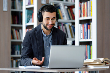 Cheerful man with eyeglasses using laptop and headphones for video call, talking to webcam, gesturing, having teleconference or zoom event, online training in library, bookcases on background - obrazy, fototapety, plakaty