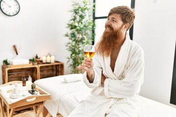 Young redhead man wearing bathrobe drinking champagne at beauty center