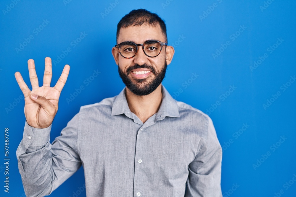 Sticker Middle east man with beard standing over blue background showing and pointing up with fingers number four while smiling confident and happy.