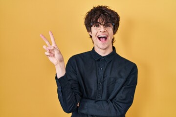 Young man wearing glasses over yellow background smiling with happy face winking at the camera doing victory sign. number two.