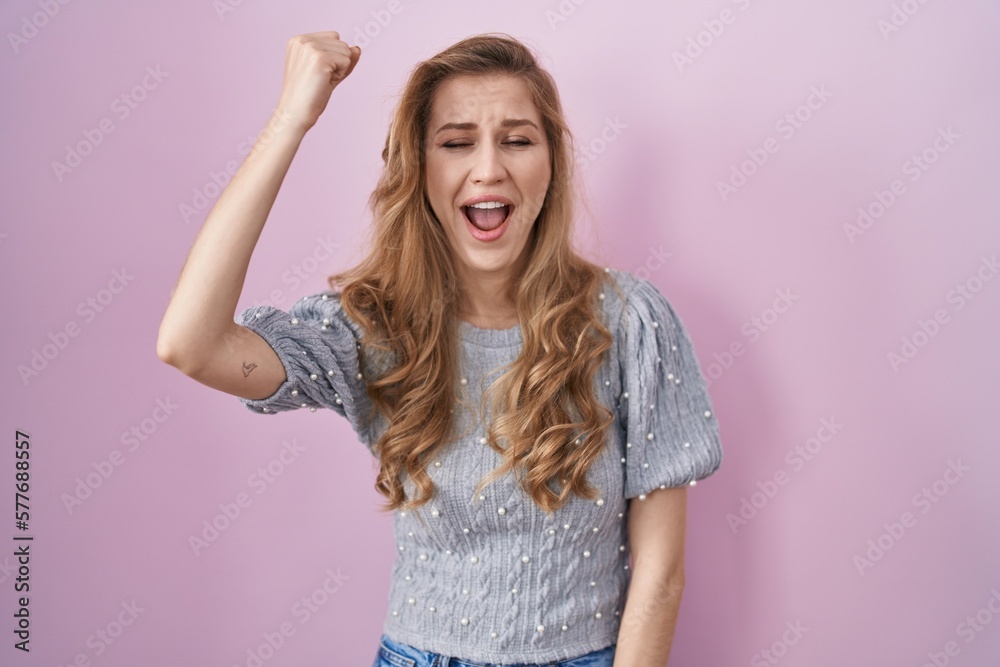 Poster Beautiful blonde woman standing over pink background angry and mad raising fist frustrated and furious while shouting with anger. rage and aggressive concept.