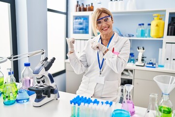 Middle age blonde woman working at scientist laboratory pointing to the back behind with hand and thumbs up, smiling confident