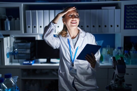 Beautiful blonde woman working at scientist laboratory late at night very happy and smiling looking far away with hand over head. searching concept.