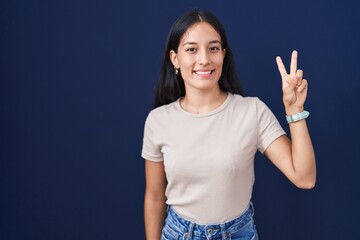 Fototapeta na wymiar Young hispanic woman standing over blue background showing and pointing up with fingers number two while smiling confident and happy.