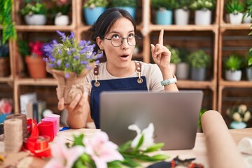 Young hispanic woman working at florist shop doing video call amazed and surprised looking up and pointing with fingers and raised arms.