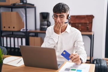 Young hispanic man working using computer laptop holding credit card laughing and embarrassed giggle covering mouth with hands, gossip and scandal concept