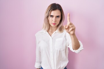 Young beautiful woman standing over pink background pointing with finger up and angry expression, showing no gesture