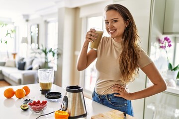 Young beautiful hispanic woman smiling confident holding glass of vegetable smoothie at the kitchen