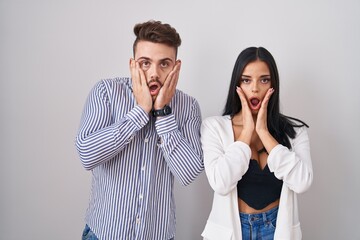 Young hispanic couple standing over white background afraid and shocked, surprise and amazed expression with hands on face