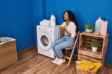 Young latin woman drinking coffee waiting for washing machine at laundru room