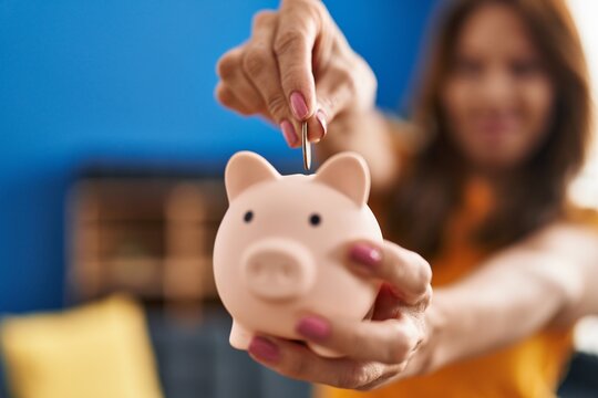 Young latin woman smiling confident insert coin on piggy bank at home