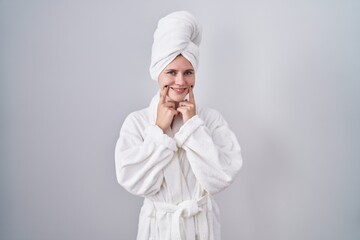 Blonde caucasian woman wearing bathrobe smiling with open mouth, fingers pointing and forcing cheerful smile