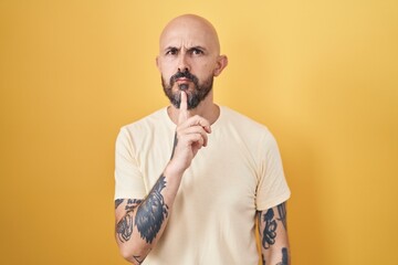 Hispanic man with tattoos standing over yellow background thinking concentrated about doubt with...