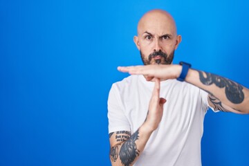 Hispanic man with tattoos standing over blue background doing time out gesture with hands, frustrated and serious face