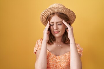 Young redhead woman standing over yellow background wearing summer hat with hand on head for pain in head because stress. suffering migraine.