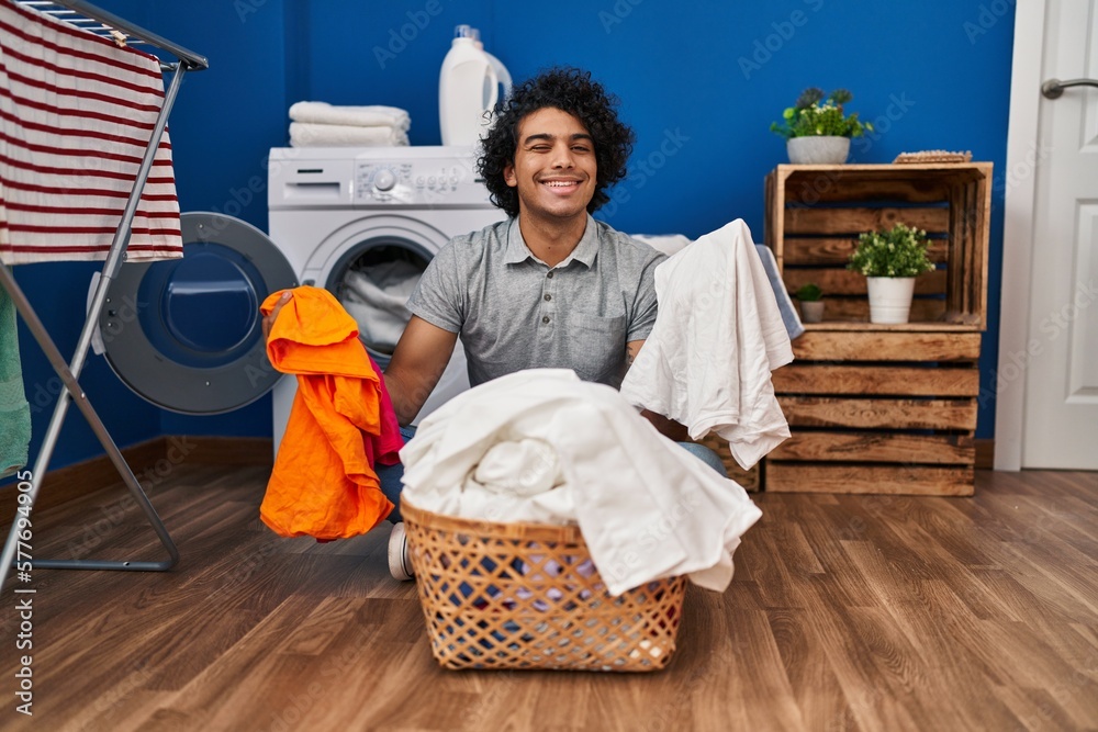 Poster Hispanic man with curly hair doing laundry at laundry room winking looking at the camera with sexy expression, cheerful and happy face.