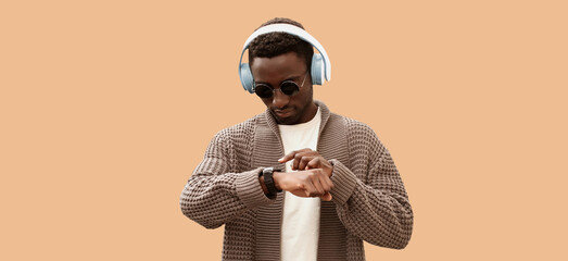 Portrait of african man listening to music in headphones looking at smart watch using voice assistant or takes calling isolated on brown background