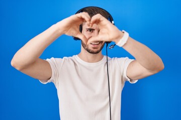 Hispanic man with beard listening to music wearing headphones doing heart shape with hand and fingers smiling looking through sign