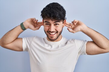 Hispanic man with beard standing over white background smiling pulling ears with fingers, funny...