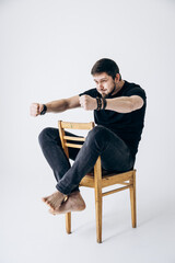 A guy in a black T-shirt sits on a chair and holds his hands like he is driving a car.