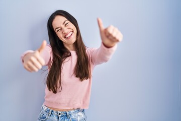 Young brunette woman standing over blue background approving doing positive gesture with hand, thumbs up smiling and happy for success. winner gesture.