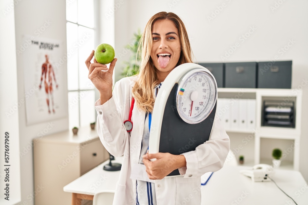 Sticker Young blonde doctor woman holding weighing machine and green apple sticking tongue out happy with funny expression.