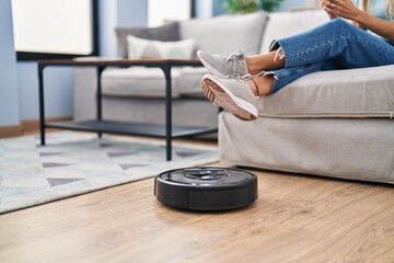 Young woman using smartphone cleaning floor using cleaner robot vacuum at home