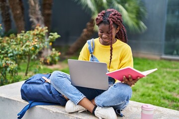 African american woman student using laptop reading book at campus park