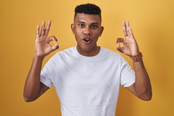 Young hispanic man standing over yellow background looking surprised and shocked doing ok approval symbol with fingers. crazy expression