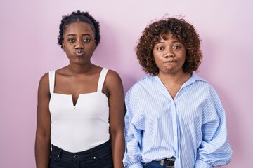Two african women standing over pink background puffing cheeks with funny face. mouth inflated with...