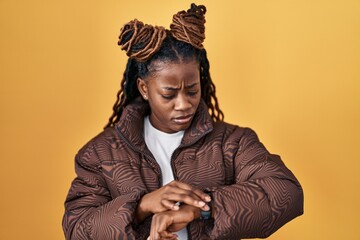 African woman with braided hair standing over yellow background checking the time on wrist watch,...
