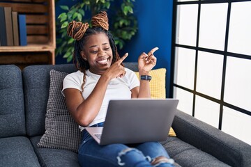 Fototapeta na wymiar African woman with braided hair using laptop at home smiling and looking at the camera pointing with two hands and fingers to the side.