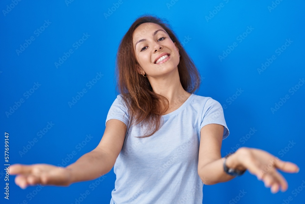 Canvas Prints Brunette woman standing over blue background smiling cheerful offering hands giving assistance and acceptance.