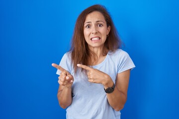 Brunette woman standing over blue background pointing aside worried and nervous with both hands,...