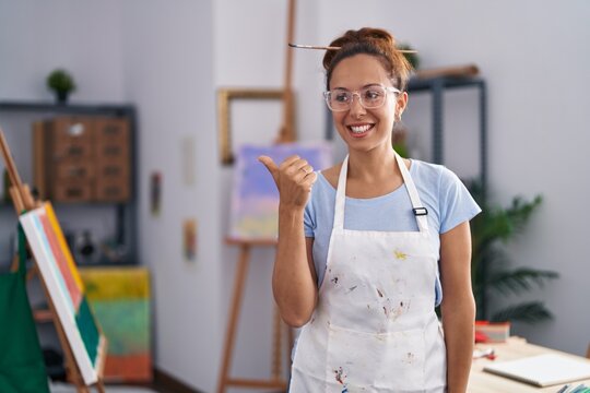 Brunette woman painting at art studio smiling with happy face looking and pointing to the side with thumb up.