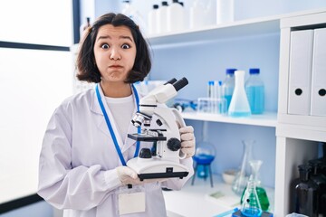 Young hispanic woman working at scientist laboratory holding microscope puffing cheeks with funny face. mouth inflated with air, catching air.