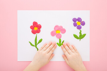 Baby girl hands modeling colorful flower shapes from clay on white paper on light pink table background. Pastel color. Point of view shot. Closeup. Toddler development. Top down view.
