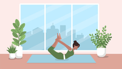 A woman in sportswear performs a yoga bow pose while lying on a mat in a room. The concept of a healthy lifestyle in a big city.