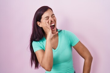Young hispanic woman standing over pink background shouting and screaming loud to side with hand on mouth. communication concept.