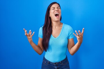 Young hispanic woman standing over blue background crazy and mad shouting and yelling with aggressive expression and arms raised. frustration concept.