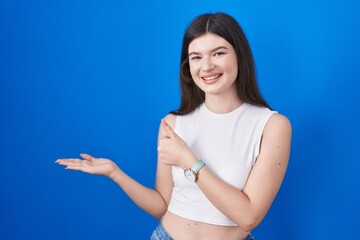 Young caucasian woman standing over blue background showing palm hand and doing ok gesture with thumbs up, smiling happy and cheerful