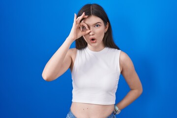 Fototapeta na wymiar Young caucasian woman standing over blue background doing ok gesture shocked with surprised face, eye looking through fingers. unbelieving expression.