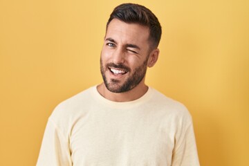 Handsome hispanic man standing over yellow background winking looking at the camera with sexy expression, cheerful and happy face.