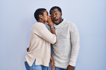 Young african american couple standing over blue background together hand on mouth telling secret...