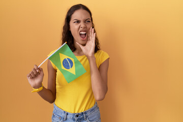 Young hispanic woman holding brazil flag shouting and screaming loud to side with hand on mouth. communication concept.