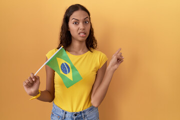 Young hispanic woman holding brazil flag pointing aside worried and nervous with forefinger, concerned and surprised expression