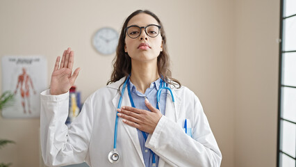 Young beautiful hispanic woman doctor making an oath with hand on chest at clinic