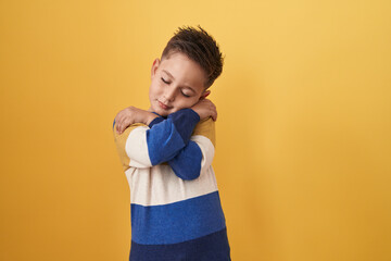 Little hispanic boy standing over yellow background hugging oneself happy and positive, smiling confident. self love and self care