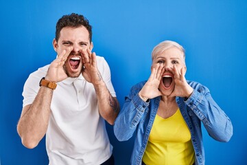 Young brazilian mother and son standing over blue background shouting angry out loud with hands over mouth